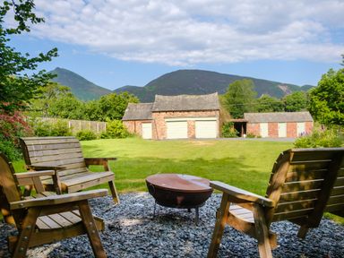 An outdoor seating area with wooden chairs and a fire pit overlooking a grassy yard and brick outbuildings with mountains in the background at Swinside Lodge in Newlands Valley near Keswick