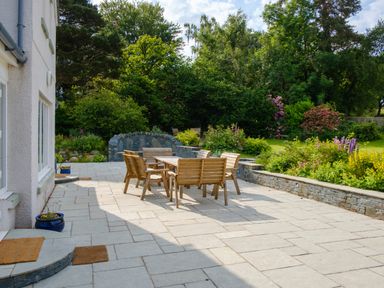 A stone patio with a wooden table and chairs next to a garden with flowers and trees at Swinside Lodge in Newlands Valley near Keswick