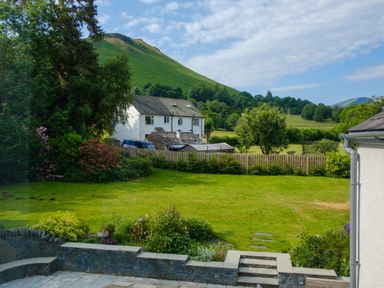 A garden with lawn shrubs and a patio area with stone steps and walls overlooking a white house and green hills at Swinside Lodge in Newlands Valley near Keswick