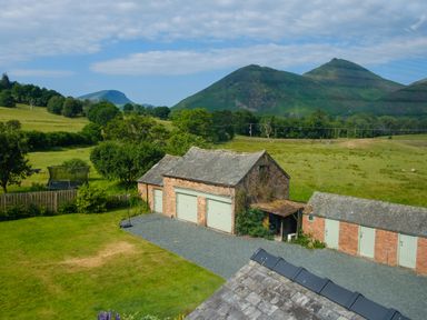 A rural scene with brick outbuildings and gravel driveway surrounded by grassy fields and hills in the background at Swinside Lodge in Newlands Valley near Keswick