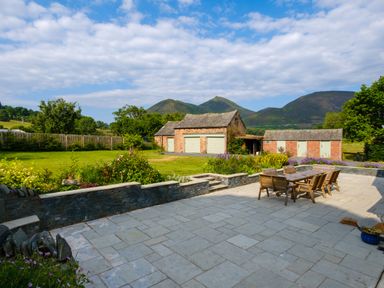An outdoor patio with table and chairs a stone wall garden area and brick outbuildings with mountains and trees in the background at Swinside Lodge in Newlands Valley near Keswick