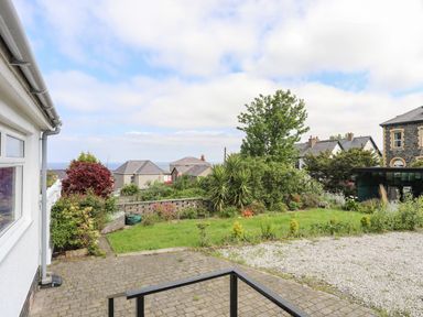 A garden with grass shrubs and trees next to a paved area and gravel driveway with houses in the background at Haddef Penmaenmawr in Penmaenmawr