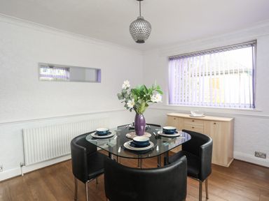A dining area with a glass table and four black chairs with tableware and a purple vase with flowers next to a window at Haddef Penmaenmawr in Penmaenmawr