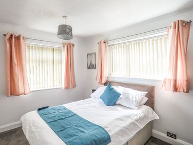 A bedroom with a double bed covered in white and blue bedding with pink curtains on two windows at Haddef Penmaenmawr in Penmaenmawr
