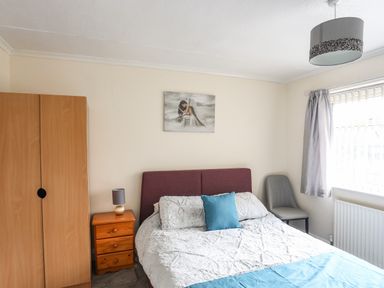 A bedroom with a bed a blue pillow wooden wardrobe bedside table with lamp and a grey chair by a window at Haddef Penmaenmawr in Penmaenmawr