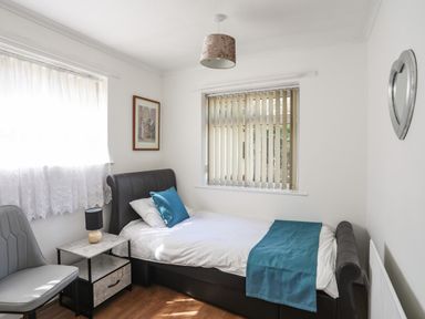 A small bedroom with a single bed a side table with a lamp a chair two windows with curtains and blinds and a heart shaped mirror on the wall at Haddef Penmaenmawr in Penmaenmawr