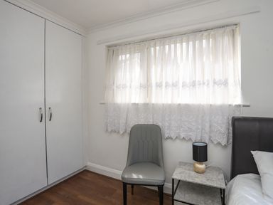 A bedroom corner with a gray chair curtain covered window a bedside table with a lamp and a bed at Haddef Penmaenmawr in Penmaenmawr