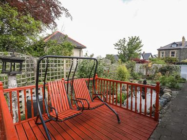 A red wooden deck with a two-seater swing overlooking a garden with a pond and trees at Haddef Penmaenmawr in Penmaenmawr