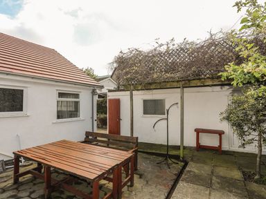 An outdoor patio area with a wooden picnic table and benches under a trellis with vines and adjacent to a white building at Haddef Penmaenmawr in Penmaenmawr