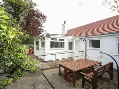 An outdoor patio area with a wooden table and bench beside a white house with red roof tiles at Haddef Penmaenmawr in Penmaenmawr