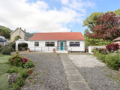 A single-story house with a red roof and a central turquoise door with a ramp and gravel driveway at Haddef Penmaenmawr in Penmaenmawr