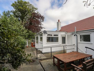 An outdoor patio area with a wooden table and chairs next to a white house with a red roof at Haddef Penmaenmawr in Penmaenmawr