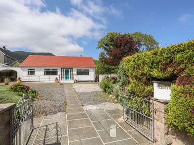 A single story house with a red roof and white walls behind a metal gate and stone driveway at Haddef Penmaenmawr in Penmaenmawr