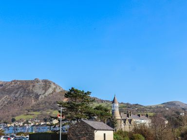 A small stone building with a conical tower near a row of houses and hills under a clear sky at Haddef Penmaenmawr in Penmaenmawr
