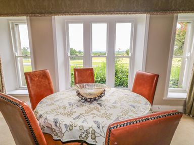 A dining area with a round table covered with a patterned tablecloth and five brown leather chairs near windows showing a garden view at Scarvagh House in Scarva near Loughbrickland