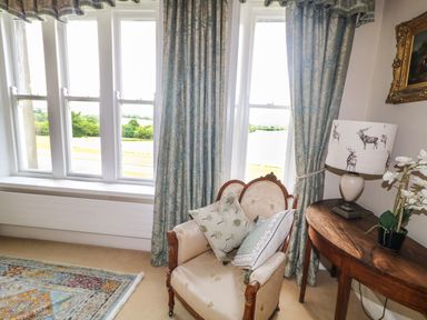 A chair with cushions near windows with curtains and a wooden table with a lamp and plant at Scarvagh House Scarva near Loughbrickland