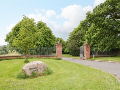 A driveway entrance with open iron gates and brick pillars near a rock with Scarvagh House engraved on it surrounded by grass and trees at Scarvagh House in Scarva near Loughbrickland
