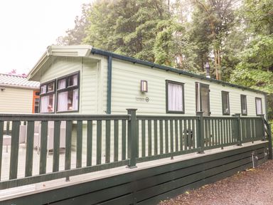 A light green static caravan with black framed windows and a dark green railing on a deck surrounded by trees at Herdwick Cabin in Troutbeck 68 White Cross Bay Holiday Park near Windermere