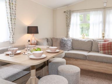 A dining area with a wooden table set with bowls and glasses surrounded by a grey upholstered corner sofa and round stools at Herdwick Cabin in Troutbeck 68 White Cross Bay Holiday Park near Windermere
