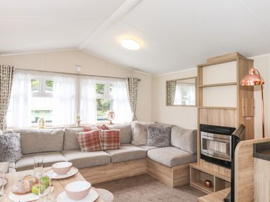 A living room with a grey sectional sofa, cushions, a fireplace, a floor lamp, and a dining table with plates and bread at Herdwick Cabin in Troutbeck 68 White Cross Bay Holiday Park near Windermere