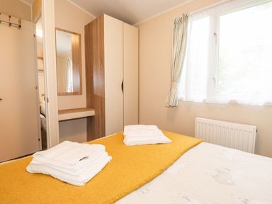 A bedroom with a bed covered with a yellow blanket and folded white towels a wardrobe and a mirror on the wall at Herdwick Cabin in Troutbeck 68 White Cross Bay Holiday Park near Windermere