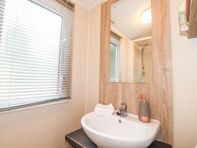 A bathroom sink with a soap dispenser and folded towels under a mirror next to a window with blinds at Herdwick Cabin Troutbeck 68 White Cross Bay Holiday Park near Windermere