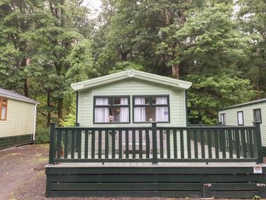 An exterior view of a light green mobile home with a dark green railing and deck surrounded by trees at Herdwick Cabin in Troutbeck 68 White Cross Bay Holiday Park near Windermere