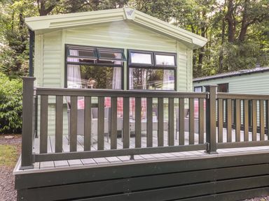 A porch with outdoor seating and a railing in front of a mobile home in a wooded area at Herdwick Cabin Troutbeck 68 White Cross Bay Holiday Park near Windermere