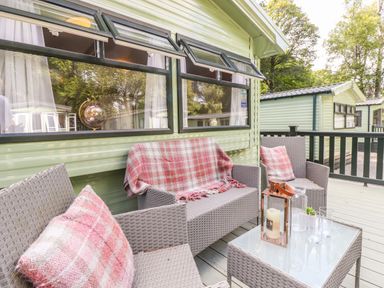 An outdoor seating area with woven chairs a sofa with plaid cushions and a glass table with a candle and glasses on a deck at Herdwick Cabin Troutbeck 68 White Cross Bay Holiday Park near Windermere
