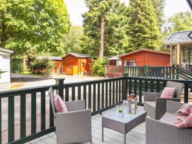 A patio with wicker chairs and table surrounded by wooden cabins and trees at Herdwick Cabin in Troutbeck 68 White Cross Bay Holiday Park near Windermere