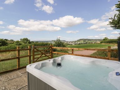 An outdoor hot tub on a stone patio next to a wooden fence with a countryside view at The Old Kennels in Harcombe near Sidford