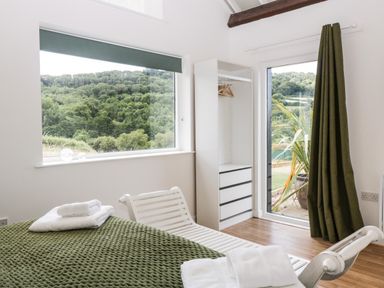 A bedroom with a green blanket on the bed and view of trees through a large window at The Old Kennels in Harcombe near Sidford