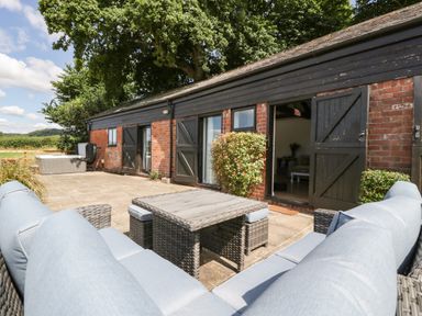 An outdoor patio with cushioned wicker seating and a table next to a brick building with black wooden doors and windows at The Old Kennels in Harcombe near Sidford
