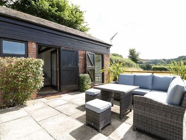 An outdoor patio with a table sofa and stools next to a building with black doors and a small hedge at The Old Kennels in Harcombe near Sidford