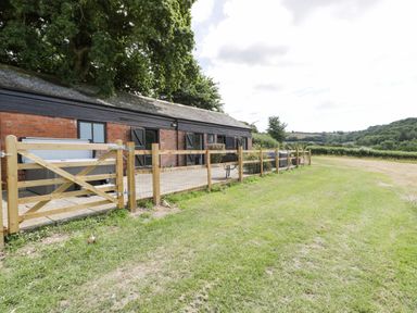 A long brick building with black shutters and a wooden gate by a grassy field at The Old Kennels in Harcombe near Sidford