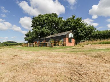 A small brick and wood building with a fenced area surrounded by grass and trees at The Old Kennels in Harcombe near Sidford