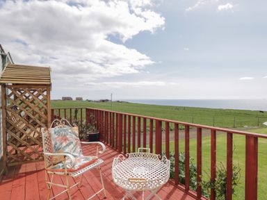 A wooden deck with two chairs and a table overlooking green fields and the sea at The Cabin in Occumster near Lybster