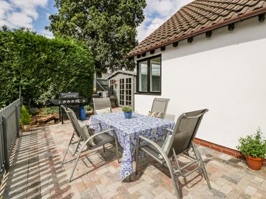 An outdoor patio area with a table covered by a blue patterned cloth and four chairs near a white house with a tiled roof and a barbecue grill at Phocle Ridge in Phocle Green near Ross-On-Wye