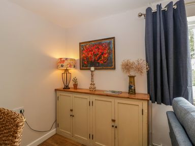 A corner of a room with a wooden cabinet topped with a floral lamp a candle holder a small plant a vase with dried flowers and a painting above blue curtains and part of a sofa visible at Phocle Ridge in Phocle Green near Ross-On-Wye