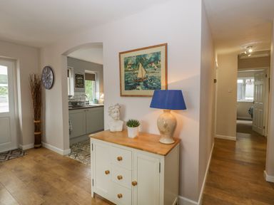 A hallway with wooden flooring a cabinet holding a lamp a sculpture and a plant a painting on the wall and an archway to a kitchen at Phocle Ridge Phocle Green near Ross-On-Wye