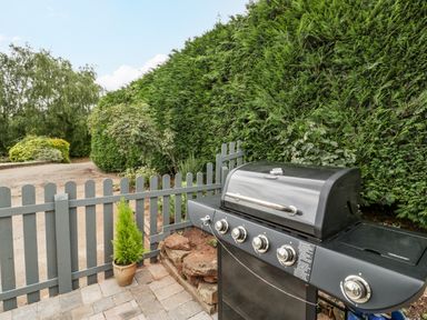 An outdoor patio area with a barbecue grill next to a grey wooden fence and tall green hedges at Phocle Ridge in Phocle Green near Ross-On-Wye