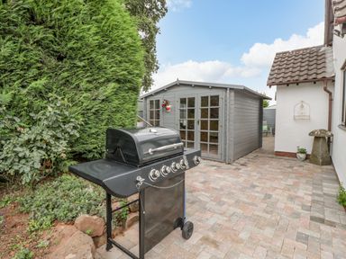 A patio with a black barbecue grill next to a grey wooden shed and white house at Phocle Ridge in Phocle Green near Ross-On-Wye