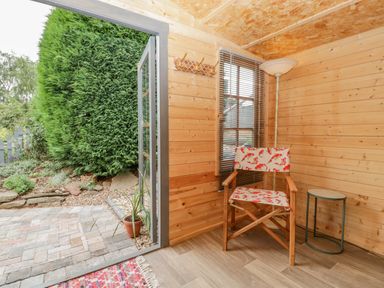 A wooden room with a chair with patterned fabric a small round table a floor lamp and an open door leading to a garden at Phocle Ridge in Phocle Green near Ross-On-Wye