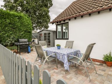An outdoor patio area with a table covered by a patterned cloth and four chairs a gray wooden shed a black barbecue grill and a gray picket fence at Phocle Ridge in Phocle Green near Ross-On-Wye