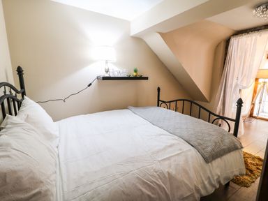A bedroom with a black metal bed frame white bedding a grey blanket a floating shelf with a lamp and decor and a window with sheer curtains at The Upper Room in Ballymena