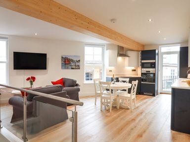 An open plan living room and kitchen with gray sofas red cushions a round dining table with four chairs and modern kitchen appliances at Ferryside Kingswear
