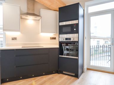 A kitchen with black lower cabinets white upper cabinets built in oven and microwave and a glass door to outside at Ferryside in Kingswear