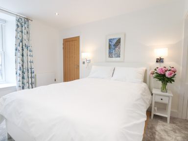 A bedroom with a white bed and pillows bedside table with flowers wooden door and patterned curtains at Ferryside Kingswear