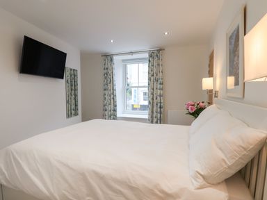 A bedroom with a white bed near a window with patterned curtains a wall-mounted TV and a vase of flowers at Ferryside in Kingswear