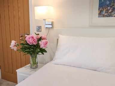 A bedroom corner with a white bed pillow a bedside table holding a vase with pink flowers a wooden door a wall light and a framed painting at Ferryside in Kingswear